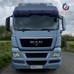 Front view of large truck on the road between two green farmlands. It is silver blue in colour