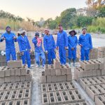 Seven workers in blue overhauls standing behind a row of building bricks inside the brick and block yard of Ngwane General Trading site