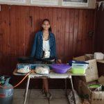 Young lady stands in front of her table on which she has a blue gas 2 plate stove and a container of samoosas which she fries