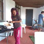 A lady with log dark hair, wearing a pink tracksuit pants, holds the microphone of the music system in front of her and behind her a young man wearing shorts and a T shirt is standing at his gas braai on the veranda of the Ifafa Beach Hotel during the Family Fun charity flea market on Saturday 28th Sept 2024